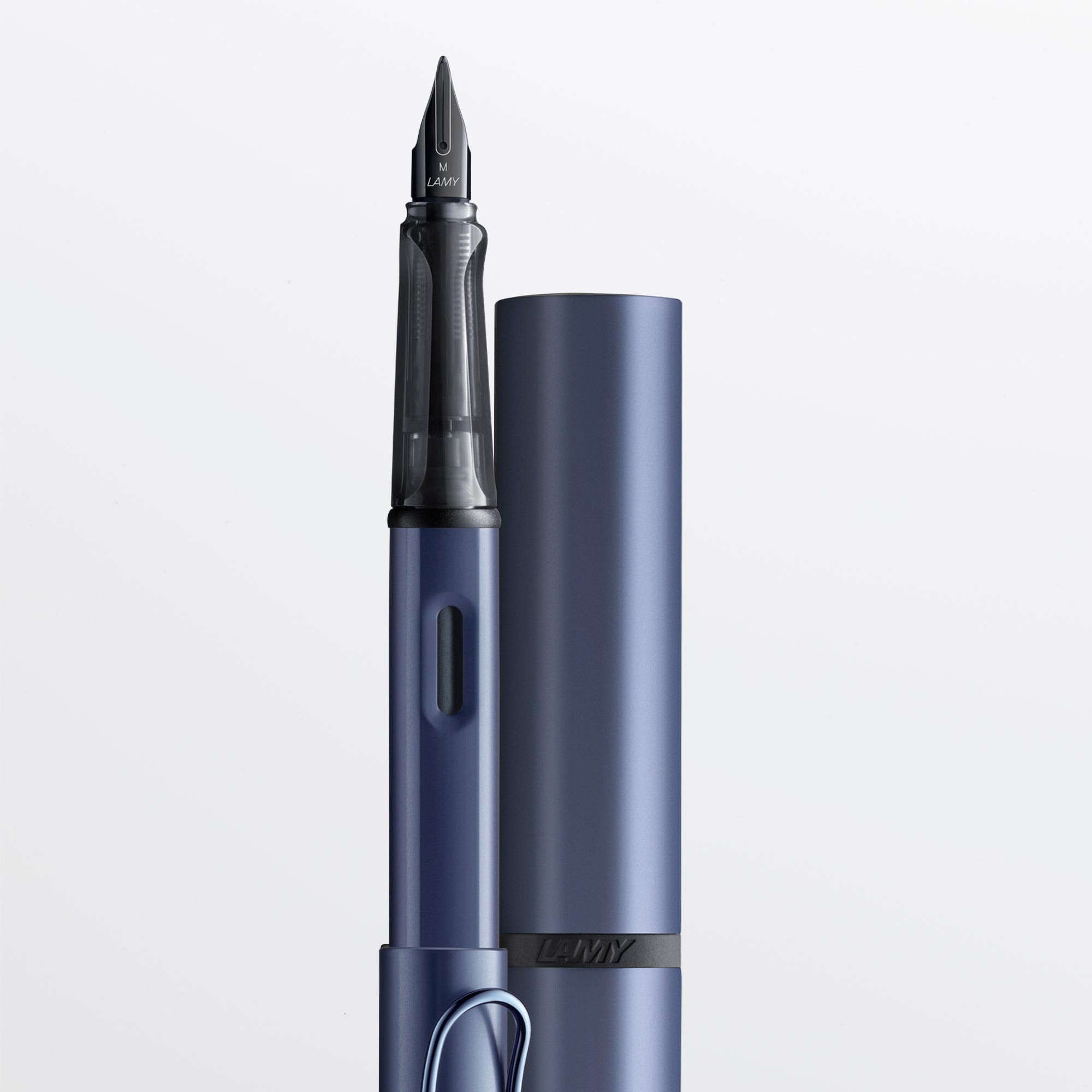 A close-up studio shot of a blue metallic fountain pen with its matching cap standing beside it on a clean white background. The nib and barrel are clearly visible, and the brand name 'LAMY' appears on the cap band and nib.
